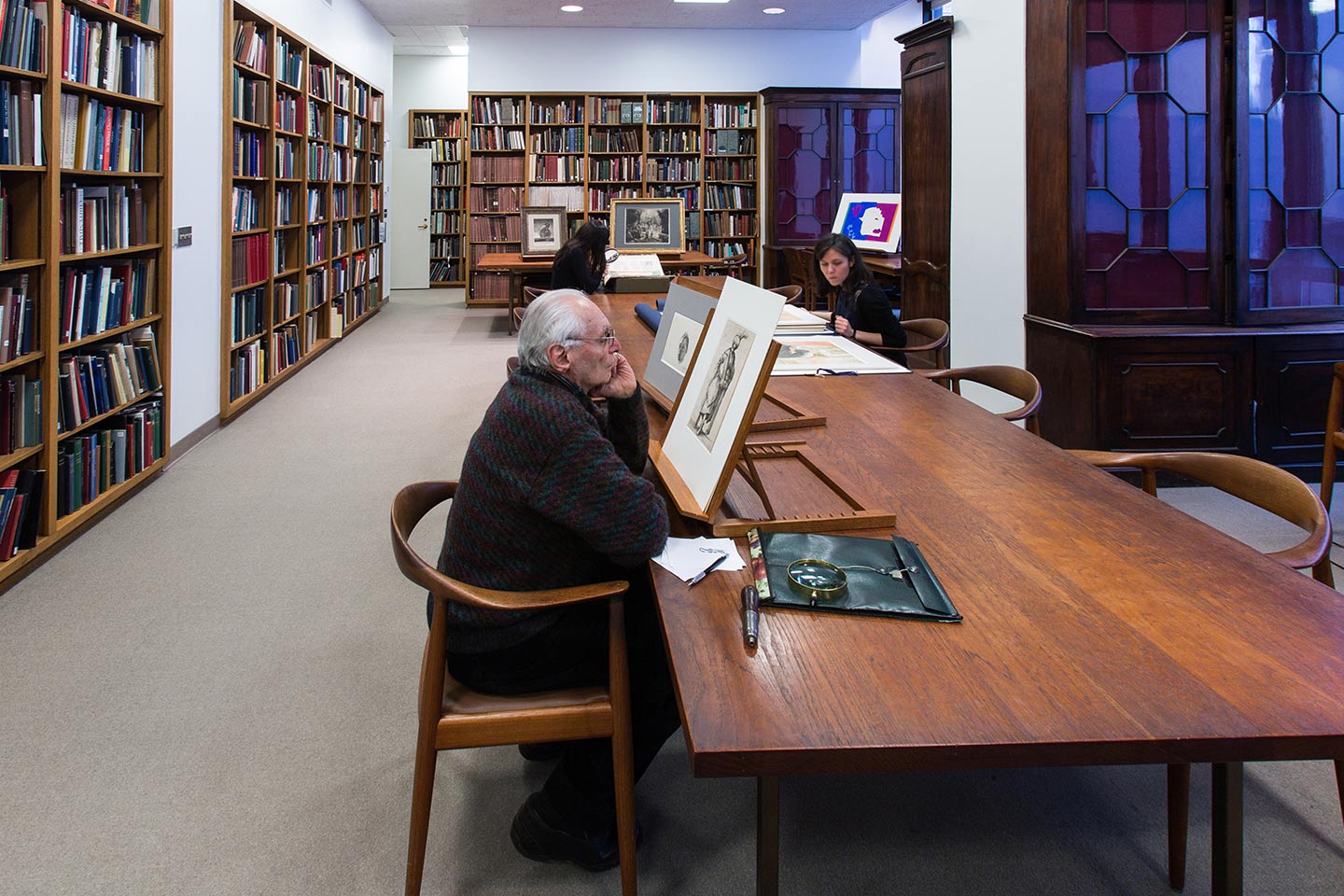 A man sits in a long wooden table staring at a black and white drawing standing before him